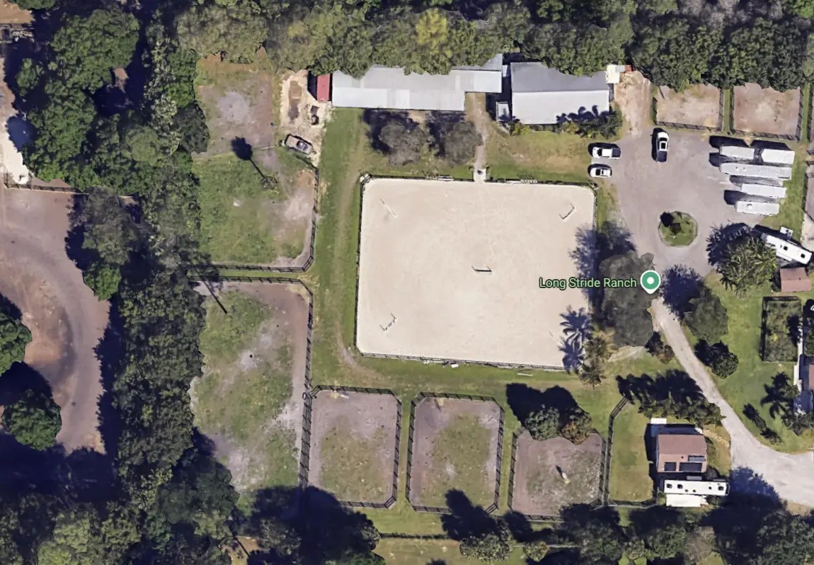 Overhead satellite view of the farm: a long covered barn along the northern edge, a large rectangular sand arena at the centre, fenced turnout paddocks to the south and west, and tree lines and sand tracks between them.