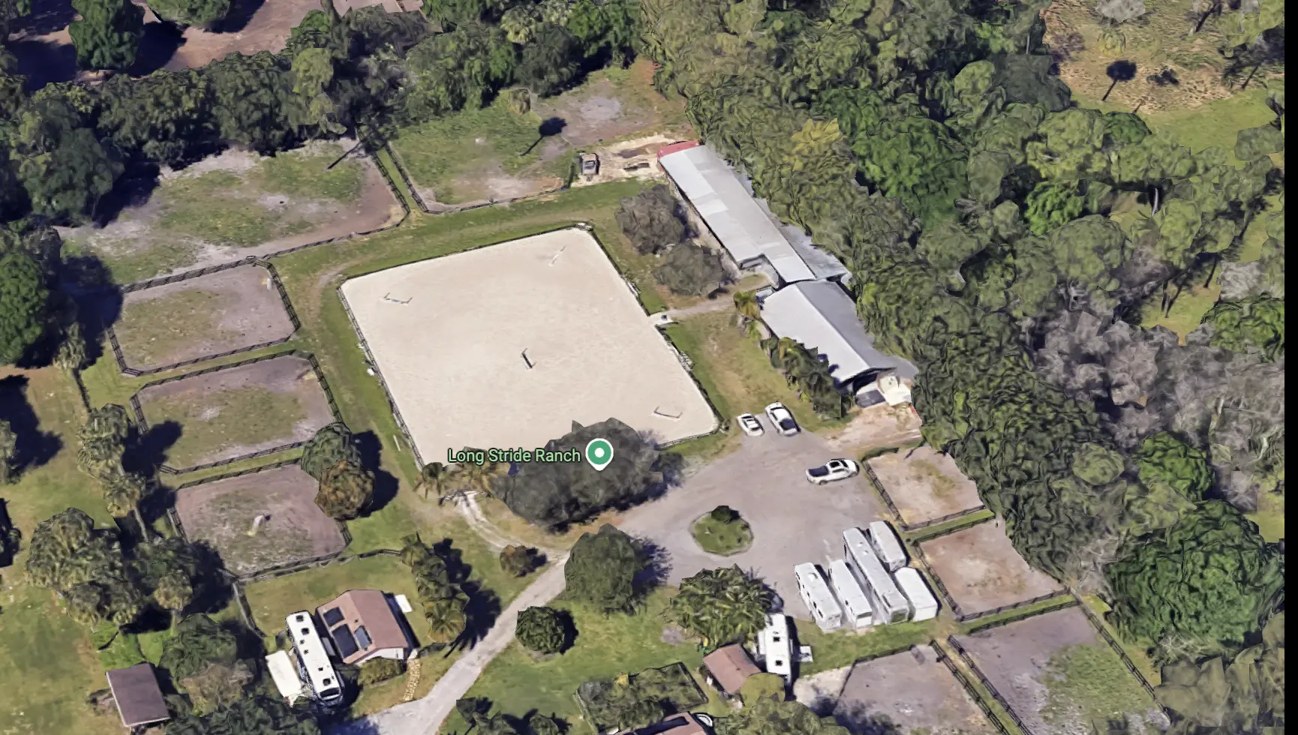 Angled aerial view of the same property, showing the arena, barn roofline, paddock fencing, house and parking areas set among mature trees.